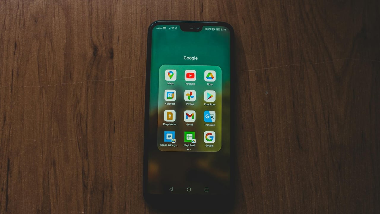 A smartphone showcasing various Google apps on a wooden table.