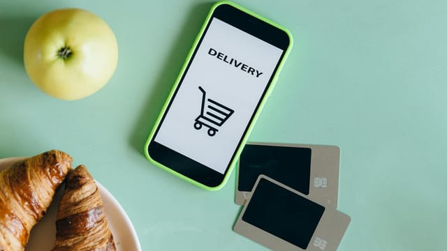 A smartphone with a delivery app next to an apple, croissants, and credit cards on a green surface.