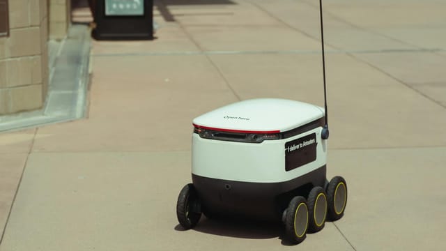 Futuristic autonomous delivery robot navigating a city sidewalk on a sunny day.