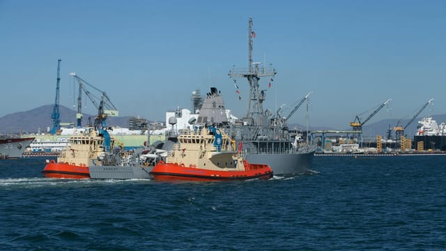 Navy vessel accompanied by tugboats in San Francisco harbor, showcasing marine industry activity.