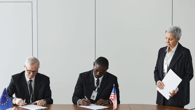 Executives signing international agreement with EU and US flags displayed on a wooden table.