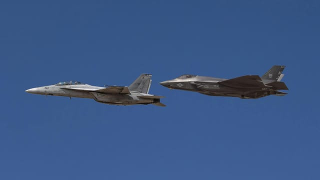 Two military jets, F-18 and F-35, flying in formation against clear blue sky.