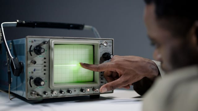 Close-up of a person pointing at a vintage oscilloscope screen in a tech setting.