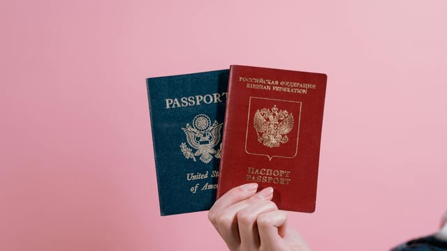 Close-up of a hand holding American and Russian passports on a pink background.