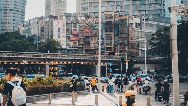 Lively city intersection with pedestrians and traffic under urban buildings featuring colorful signage.