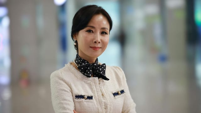 A sophisticated woman smiles confidently in a stylish outfit indoors, exuding elegance and charm.