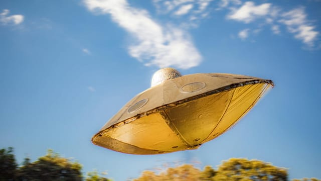 Close-up of a metallic UFO model flying against a clear blue sky background.