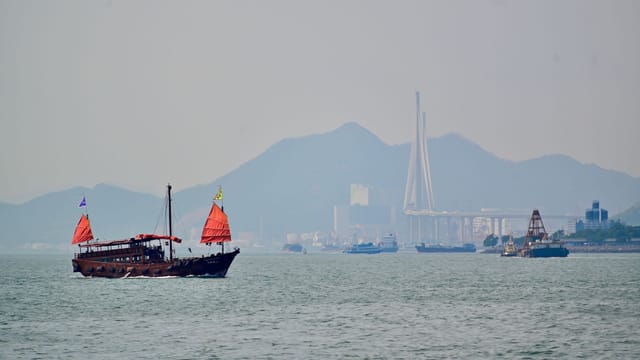 A traditional junk boat with red sails sails past a modern coastal cityscape and bridge.