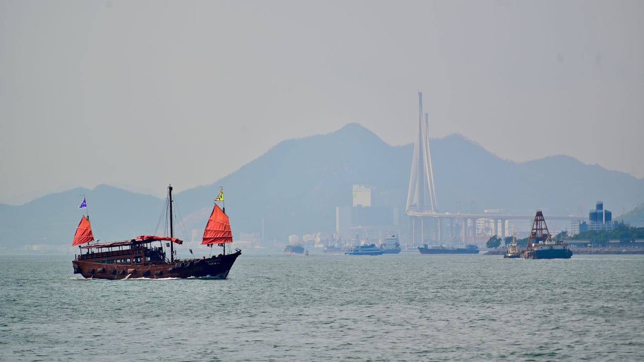 A traditional junk boat with red sails sails past a modern coastal cityscape and bridge.