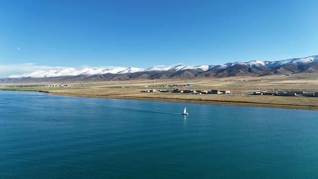 A tranquil view of Qinghai Lake with a sailboat and snow-capped mountains in the distance under a clear blue sky.