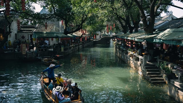 Peaceful boat ride along a canal with traditional market stalls and lanterns, under a canopy of trees.