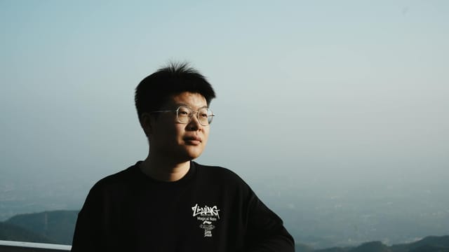 A man with glasses stands on a balcony, gazing at the mountain view under a clear blue sky.