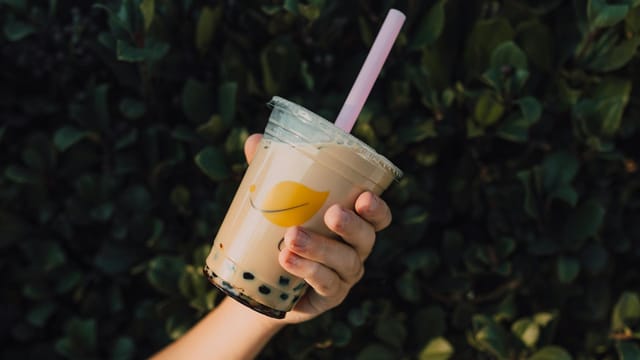 Close-up of a hand holding a refreshing milk tea with boba, outdoors against greenery.