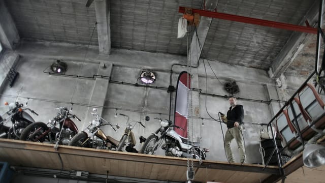 From below of bearded mechanic in casual outfit standing near motorcycles on special platform in garage and holding control panel in hand
