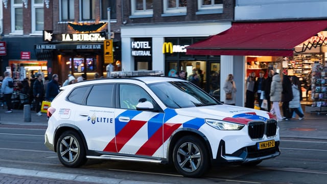 A Dutch police car on a busy street in Amsterdam with shops and pedestrians.