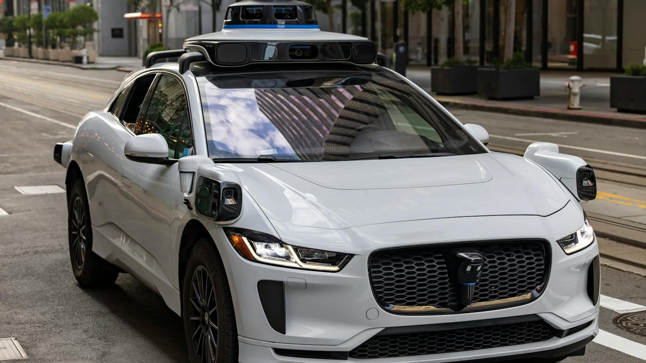 A white autonomous vehicle navigating a city street, reflecting urban architecture in daylight.