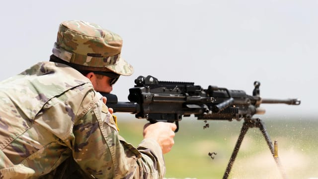 A soldier in camouflage uniform fires an M249 machine gun in an outdoor setting.