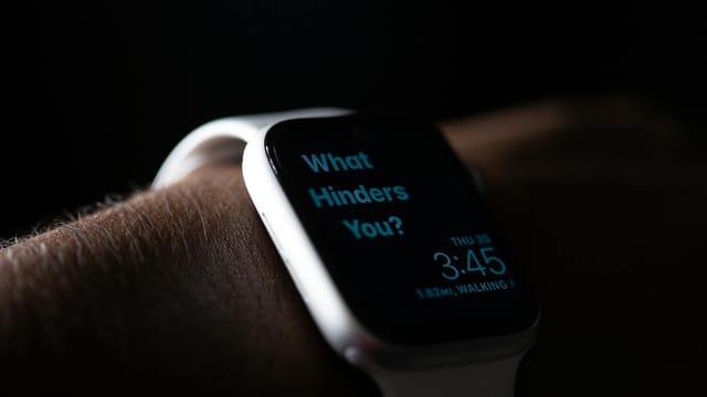 Close-up of a smartwatch showing the text 'What Hinders You?' in a dim setting.