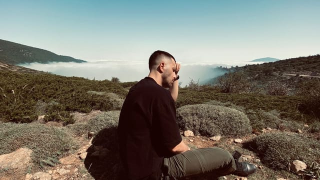 A man sits thoughtfully in a mountainous landscape with a distant view and clear sky.