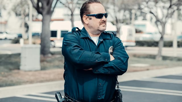 A police officer in uniform stands confidently outdoors with arms crossed and sunglasses.