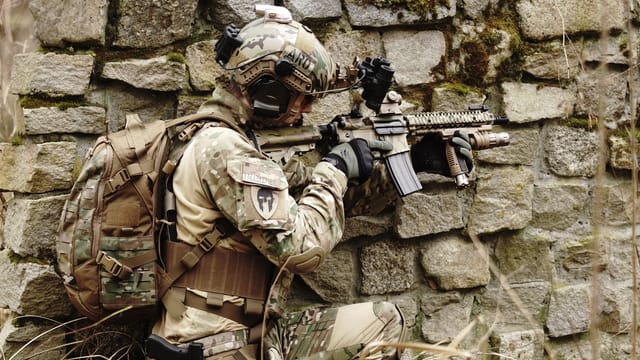 Illustrative image: Soldier in camouflage gear aims a rifle while hiding along a stone wall outdoors.