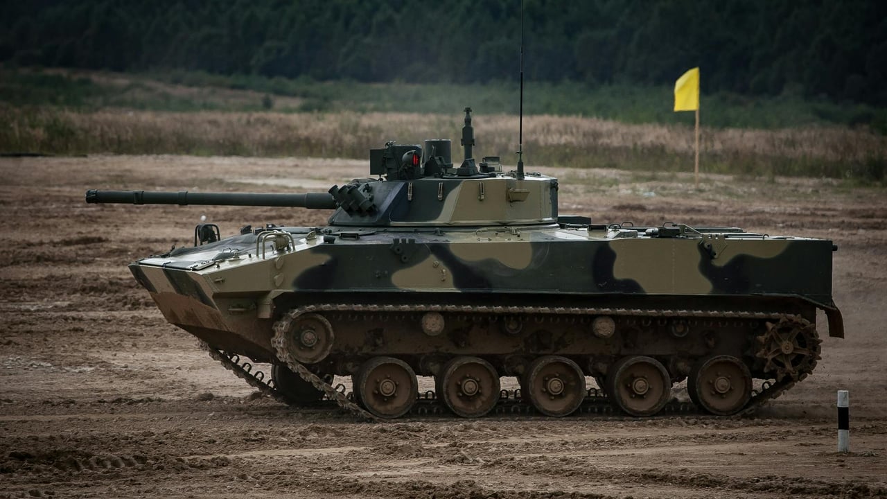 A camouflaged tank driving through a muddy field with trees in the background.