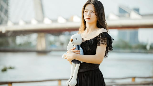 Woman in black dress holds teddy bear by the river with bridge in background during daytime.