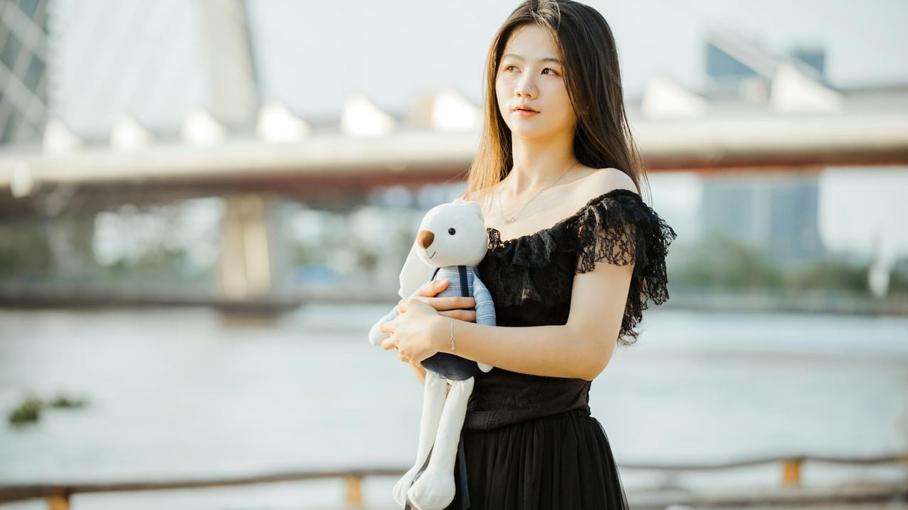 Woman in black dress holds teddy bear by the river with bridge in background during daytime.