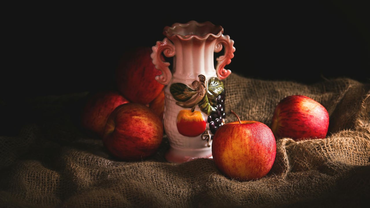 Beautiful still life of red apples and ornate ceramic vase on burlap in low light.