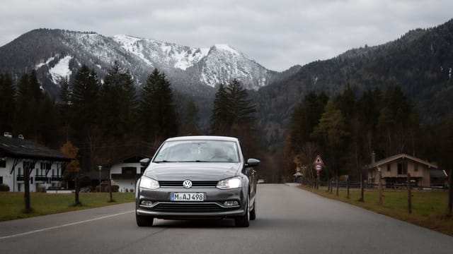 A car travels down a picturesque road near Tegernsee, Germany, with snowy mountains in the background.