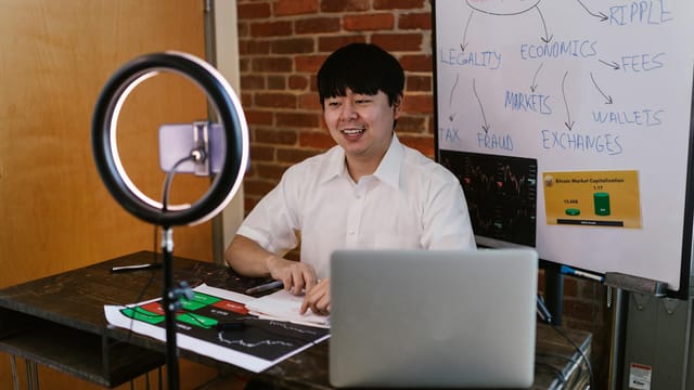 Man discussing cryptocurrency economics with ring light and laptop, engaging content creation.