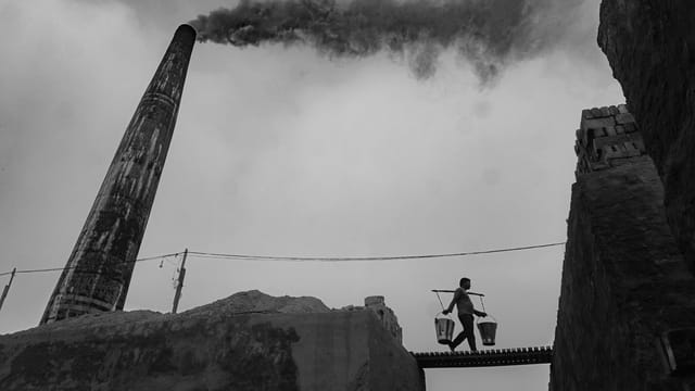 A worker carries buckets across a narrow bridge by a towering smokestack emitting black smoke, creating a dramatic industrial scene.