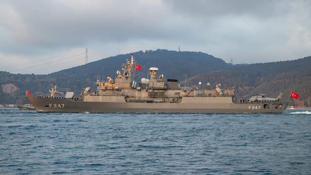Turkish warship F247 cruising through calm waters with a scenic mountainous backdrop.