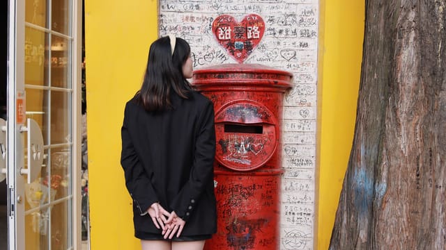 A young woman stands by a vibrant red postbox against a yellow wall in Nanjing, China.