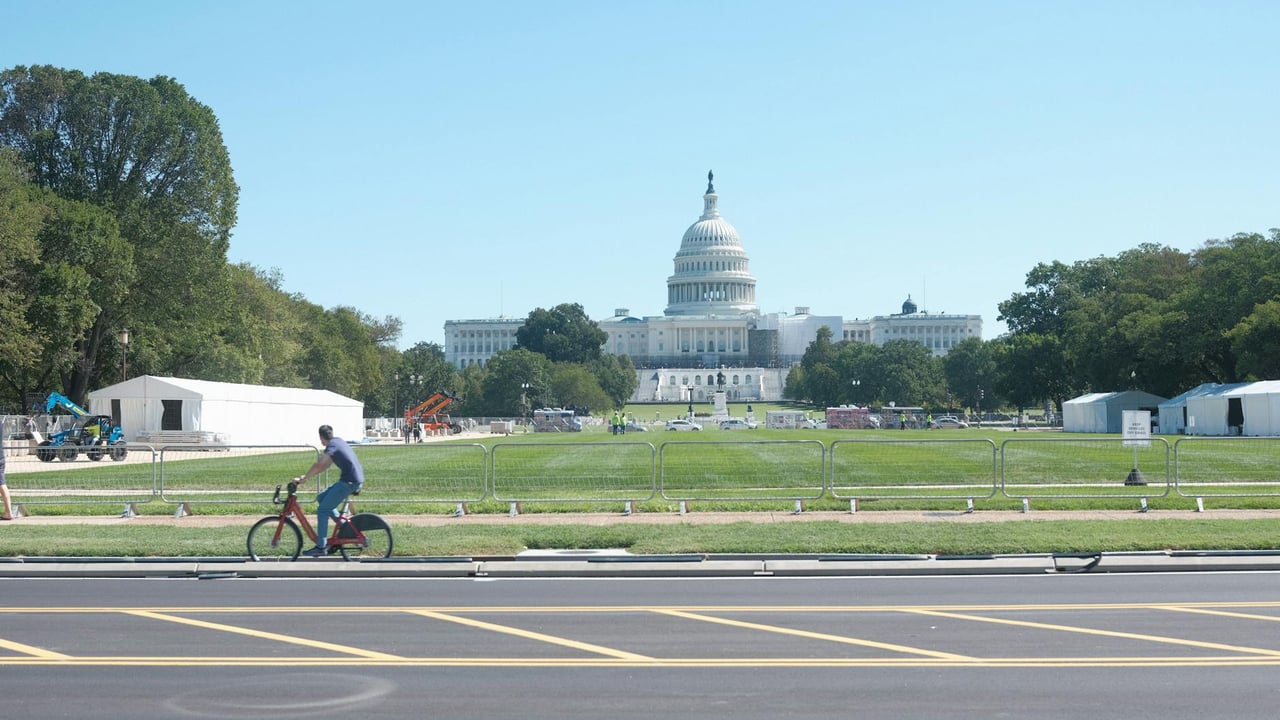 A cyclist rides past the iconic US Capitol in sunny Washington DC, surrounded by trees and open lawn.