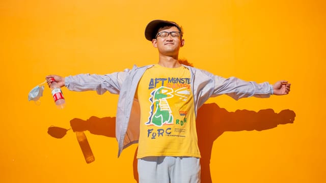 Asian man soaking up sunshine with arms outstretched, wearing casual attire and glasses.