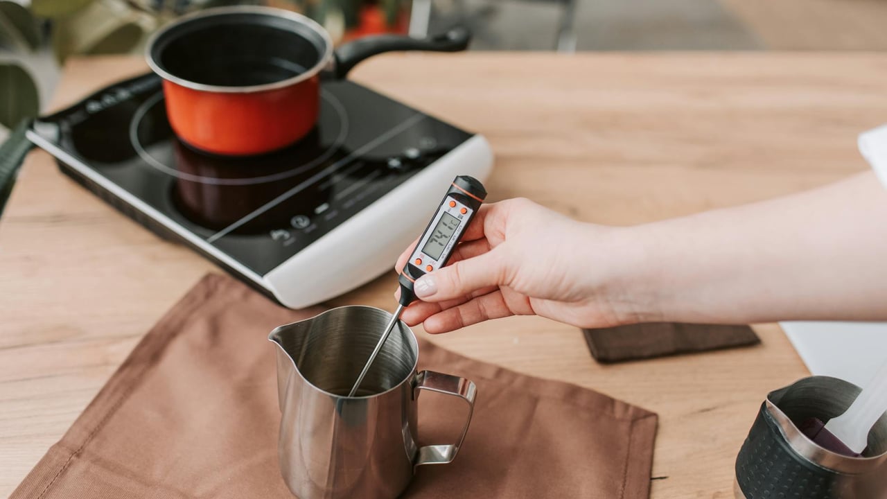 A person measures the temperature of melted wax on an electric stove, highlighting modern craft techniques.