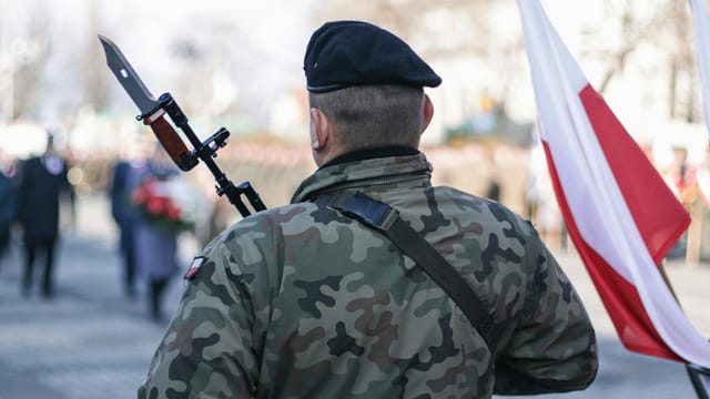 Back view of a Polish soldier at a military ceremony, holding a weapon next to the national flag.