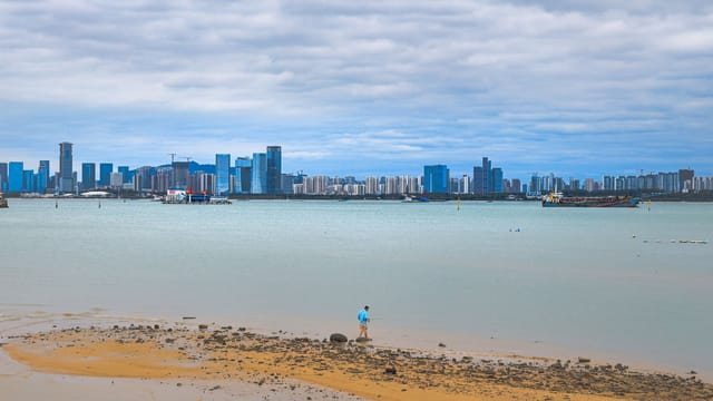 A distant skyline of Haikou, China viewed from a beach, with one person walking on the sand.