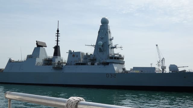 A sleek naval destroyer anchored in Portsmouth harbor, England, under a bright sky.