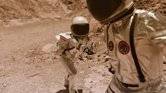 Two astronauts in space suits help each other in a desert-like terrain, evoking Mars exploration.