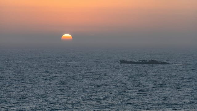 A solitary cargo ship sails under a vibrant sunset sky over the expansive ocean waters.