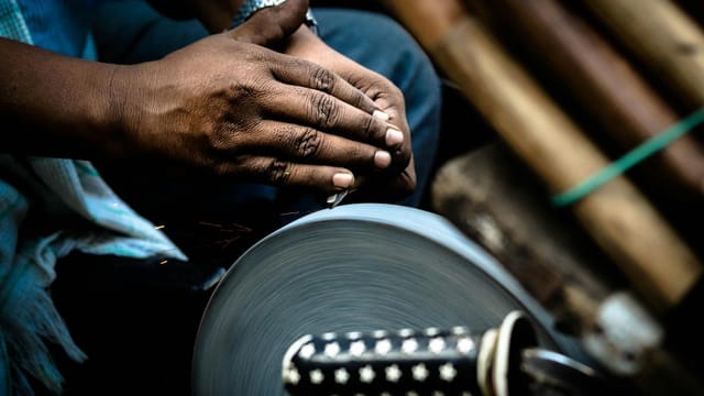 Hands sharpening a knife against a rotating grindstone in a market setting.
