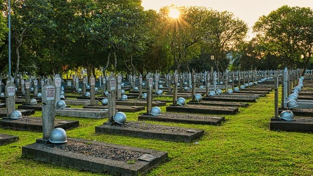 Rows of tombstones with military helmets located on grassy ground near tall lush green trees in heroes cemetery in Kalibata