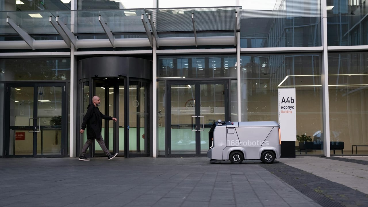 A modern office setting featuring a man and an autonomous delivery robot outside a glass-covered building.