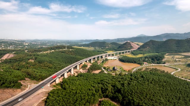 A picturesque highway stretches across lush green mountains under a bright blue sky.
