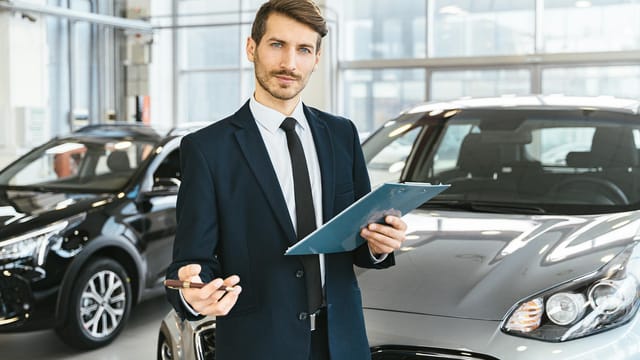 Professional car dealer in business suit holding clipboard in a bright car showroom.