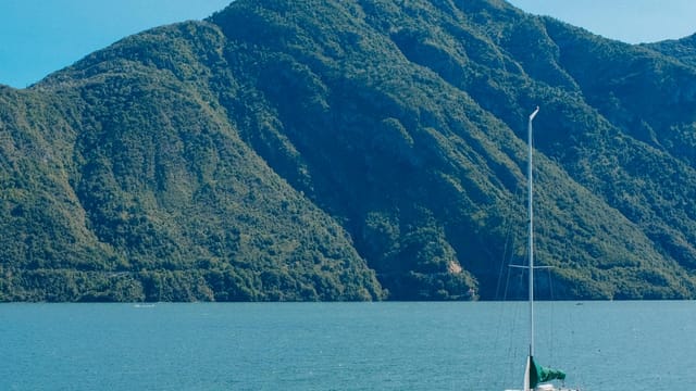 Tranquil lake with a sailboat surrounded by lush green mountains under a clear blue sky.