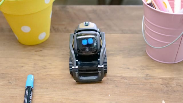 A small modern robot placed on a wooden desk surrounded by office supplies, showcases innovative technology.