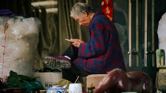 Elderly woman in traditional jacket, seated at outdoor market, browsing smartphone.
